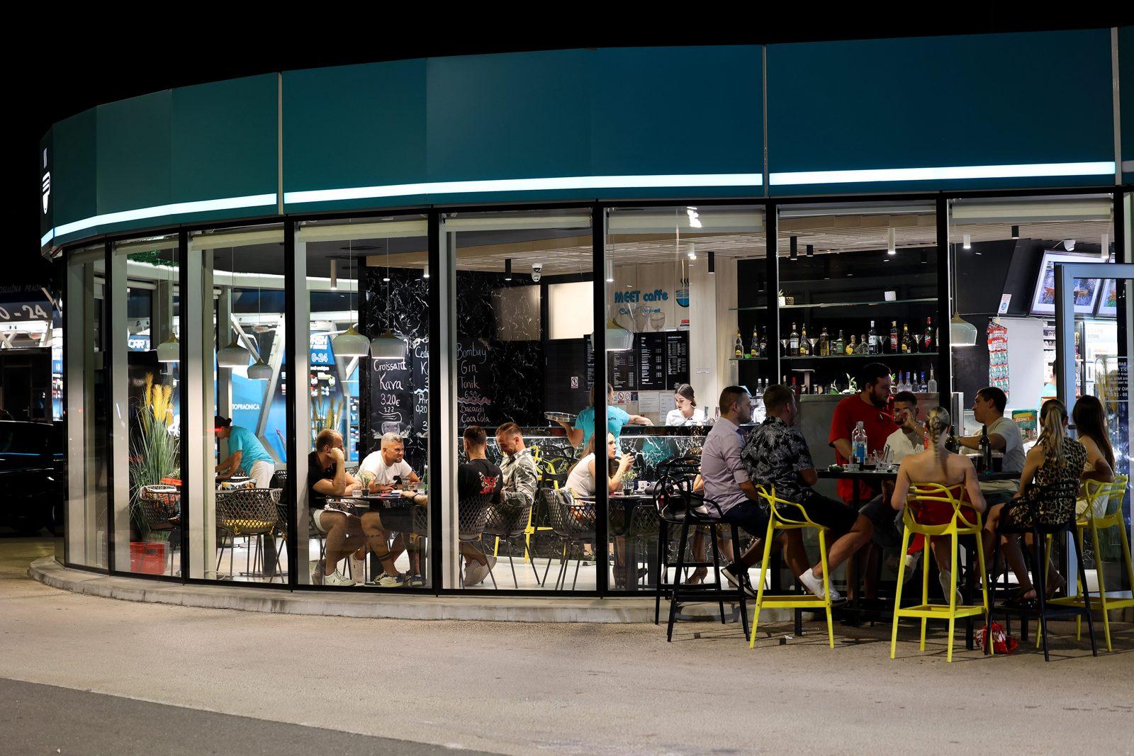 Guests at the bar of the Meet Caffe at Adria Petrol seen through the glass front at night
