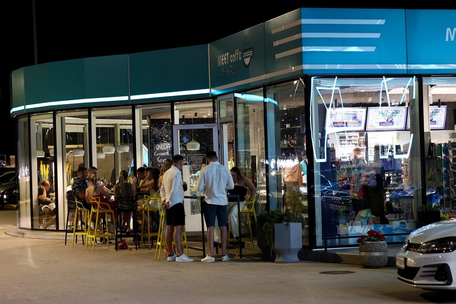 The glass front of the Meet Caffe at Adria Petrol in the evening with guests on the terrace