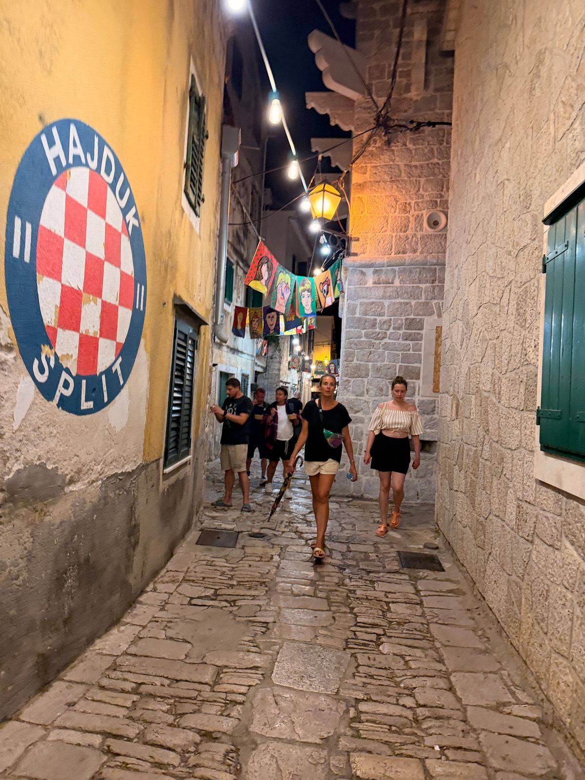 Kopfsteingasse in Vodice, zwischen den Häusern hängen Gemälde an einer Lichterkette
