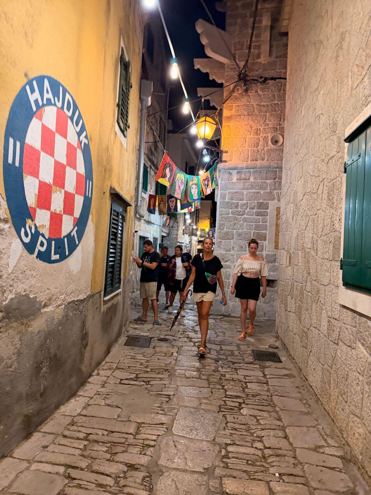 Kopfsteingasse in Vodice, zwischen den Häusern hängen Gemälde an einer Lichterkette