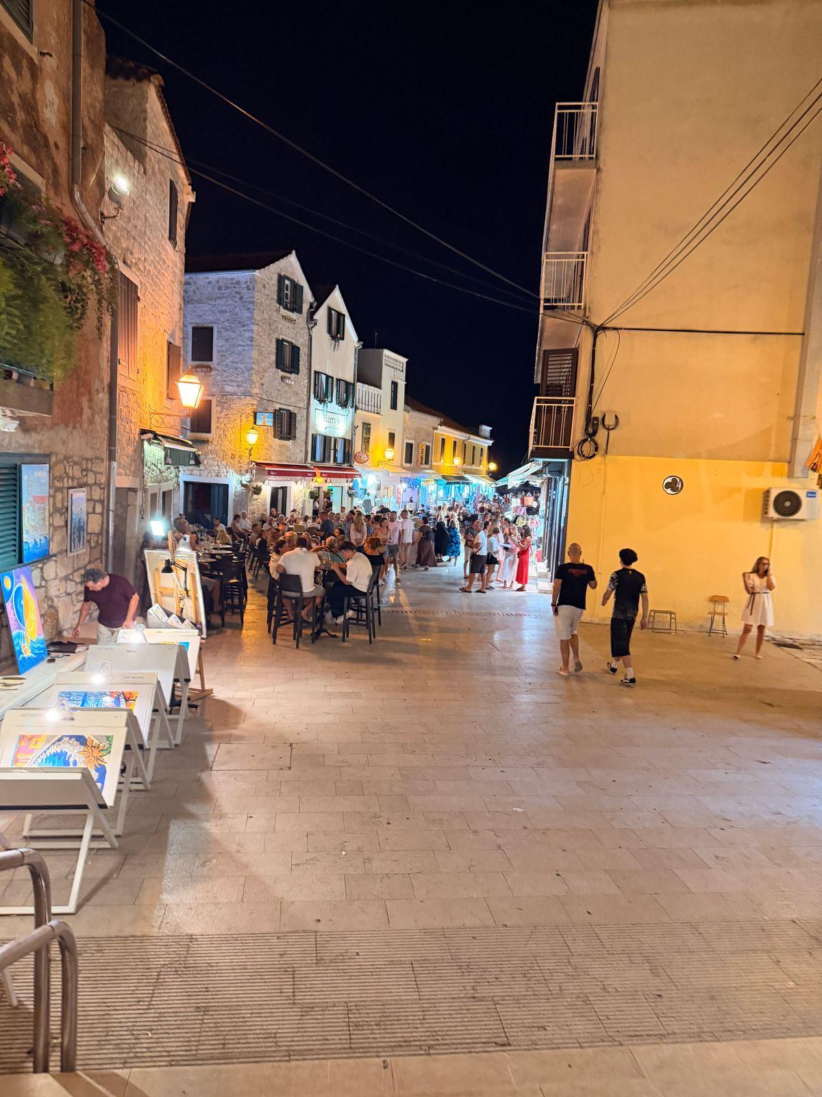 Abendliche Gasse in Vodice mit den Bildern eines Straßenmalers und Restauranttischen